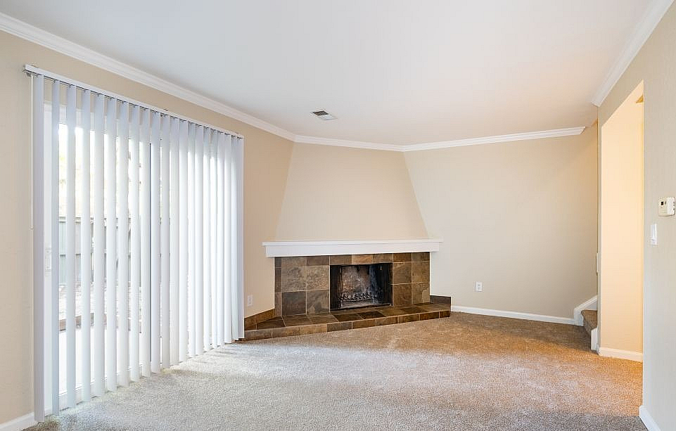 Bright living room featuring a cozy fireplace, new carpeting, and large windows with vertical blinds, offering a warm and inviting atmosphere.