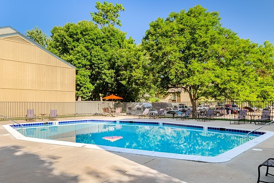 Inviting apartment complex pool area surrounded by lush trees, featuring comfortable lounge chairs and vibrant orange umbrellas under a clear blue sky.