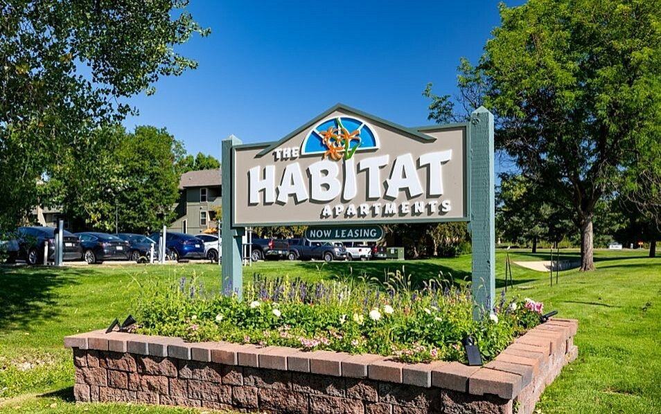 Welcoming entrance sign of The Habitat Apartments surrounded by lush greenery and vibrant flowers, now leasing.