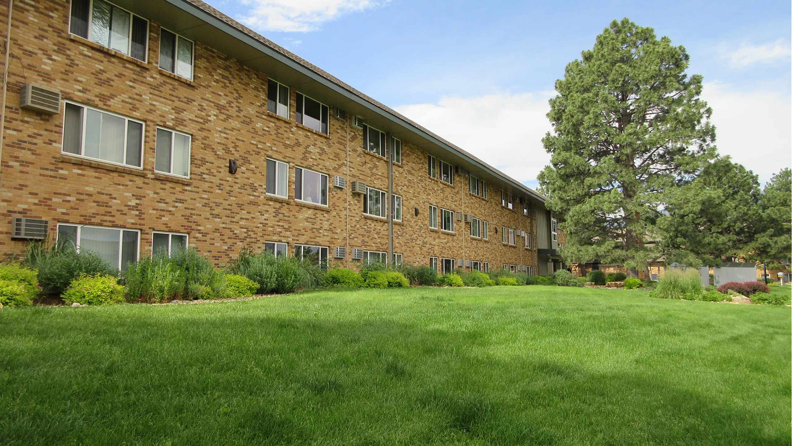 Charming brick apartment building with lush green landscaping and serene trees, set against a clear blue sky.