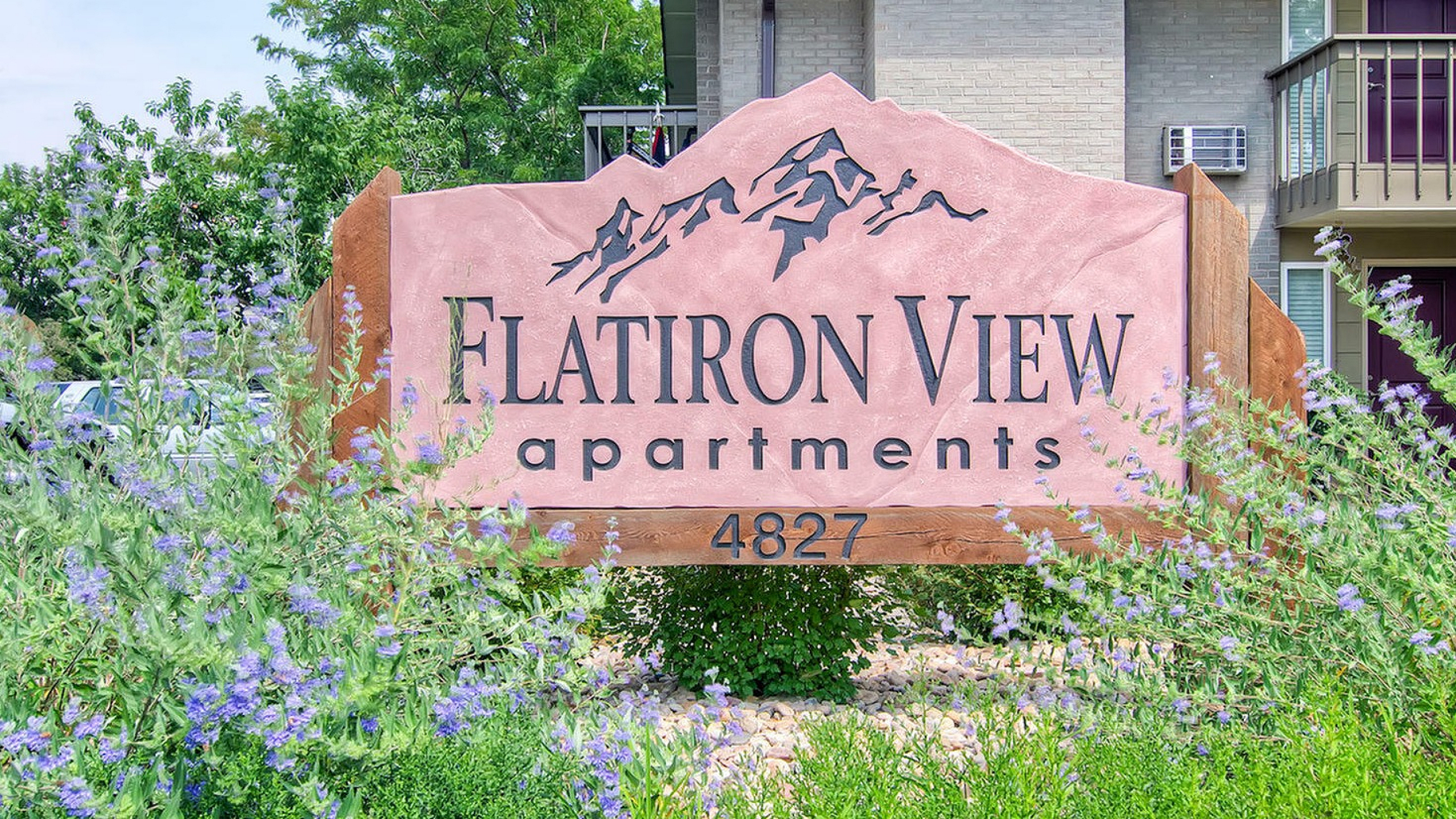 Inviting entrance sign for Flatiron View Apartments, surrounded by lush greenery and vibrant purple flowers.