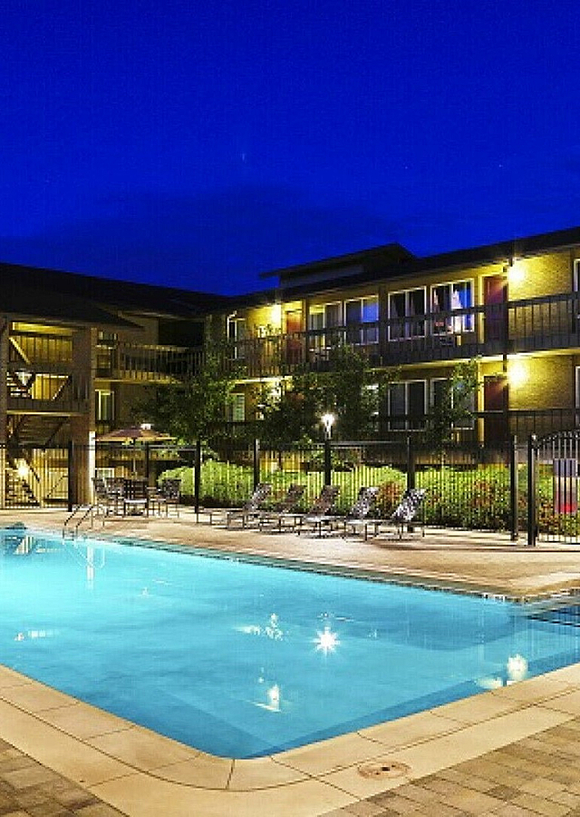 Luxurious apartment complex with a beautifully illuminated swimming pool, surrounded by lush greenery and modern architecture under a stunning twilight sky.