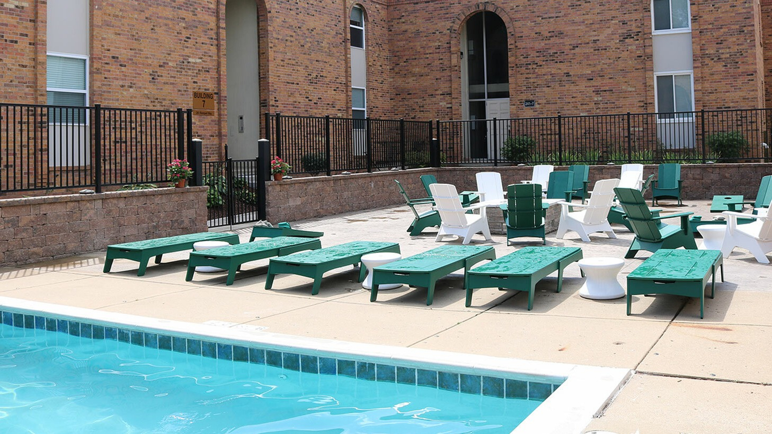 Charming brick apartment building with an inviting outdoor pool area featuring green and white lounge chairs, perfect for relaxation and leisure.