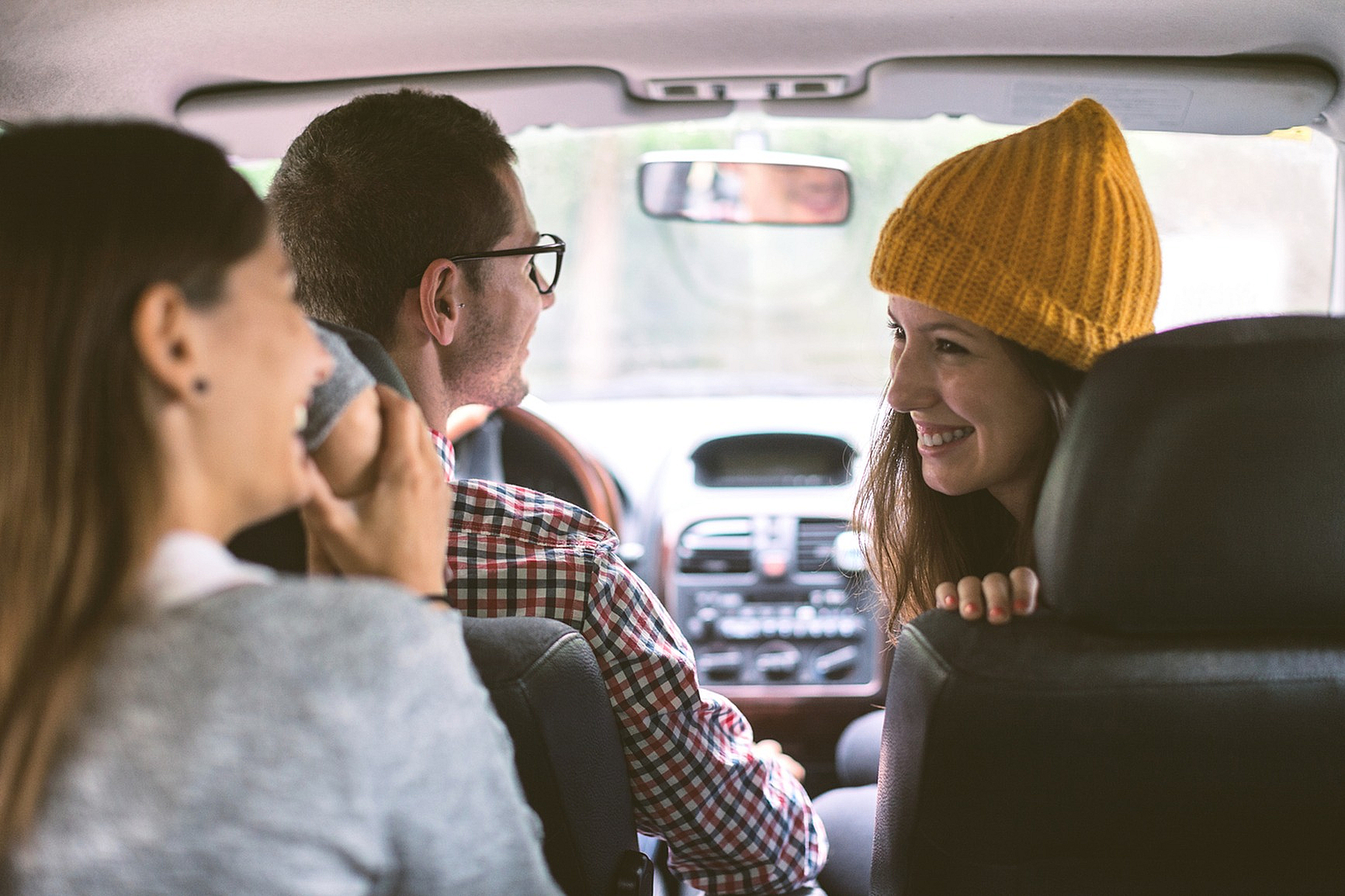 Three friends laughing and talking during a car ride, with one wearing a yellow knit hat.
