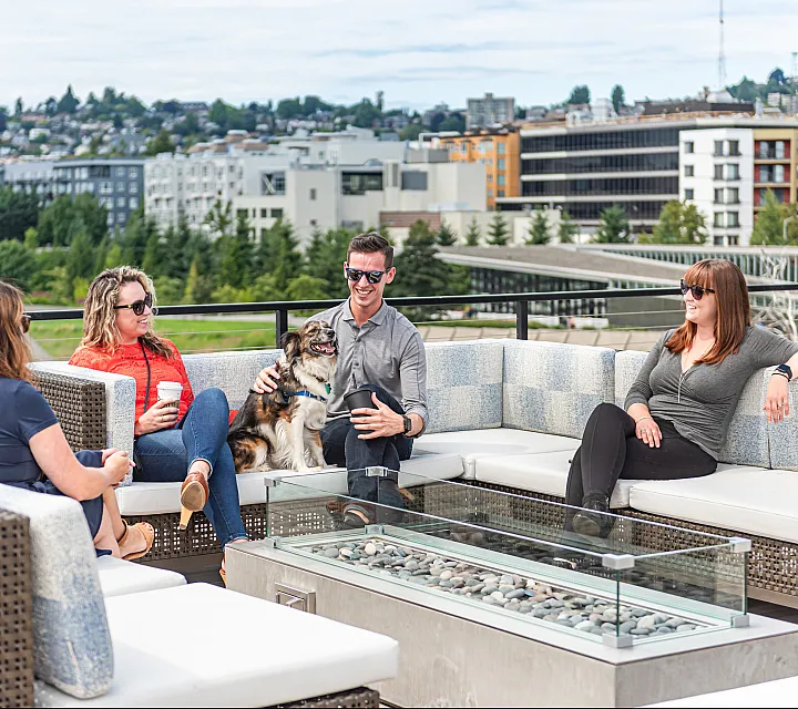 A group of four friends relax on a stylish rooftop lounge with a modern fire pit, enjoying the city view. A man in sunglasses pets a happy dog while chatting, as others sit comfortably with drinks in hand. The background features a cityscap