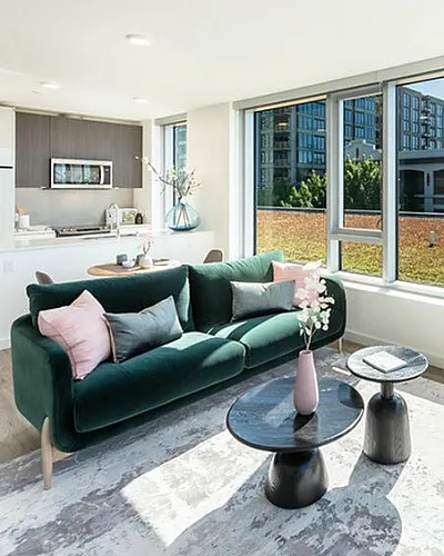 A bright, modern apartment living room with a green velvet sofa adorned with pink and gray throw pillows. A set of contemporary black coffee tables sits on a textured area rug, with a pink vase and flowers as decor. Large windows allow natu