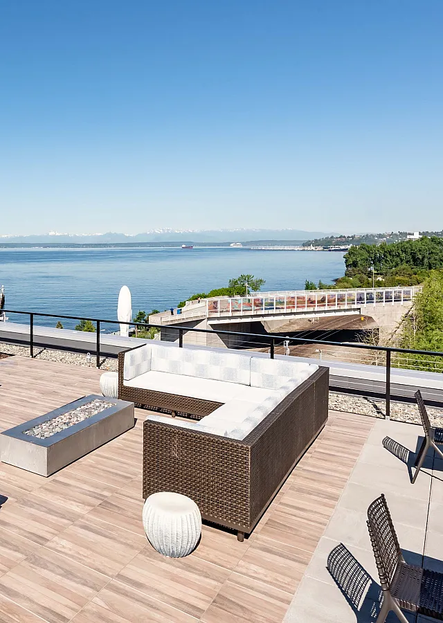 A modern rooftop lounge featuring an L-shaped wicker sofa with white cushions, a sleek fire pit, and wooden decking. The space overlooks a scenic waterfront with a clear blue sky, lush greenery, and distant mountains, framed by a black meta
