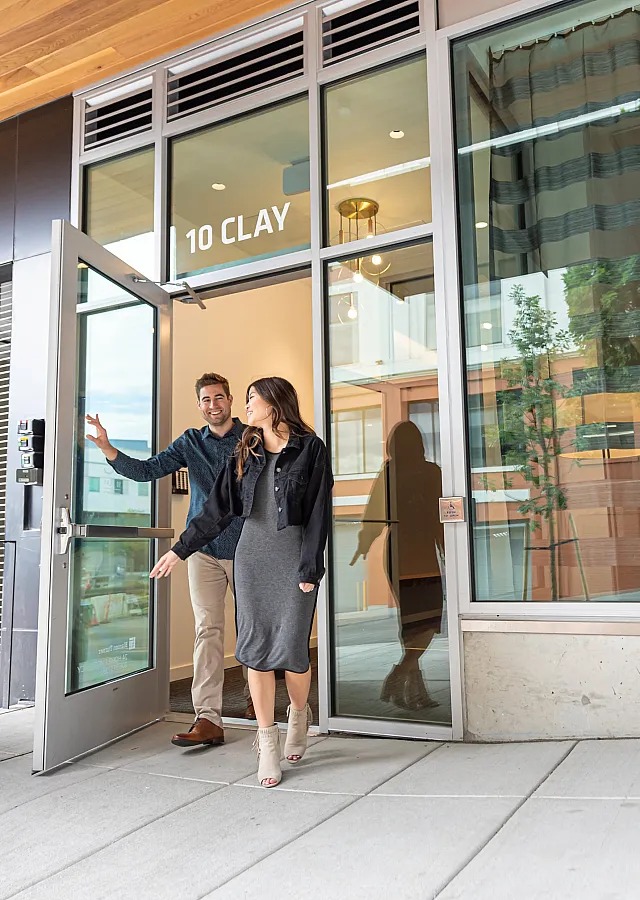 A stylish couple exits a modern apartment building named '10 Clay' through a glass door. The man, wearing a dark sweater and beige pants, holds the door open with a smile, while the woman, dressed in a gray dress and black jacket,