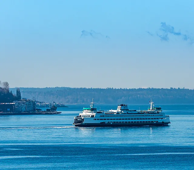 A Washington State ferry sails across calm blue waters with a scenic shoreline and tree-covered hills in the background. The sky is clear with a few wispy clouds, creating a peaceful maritime scene.