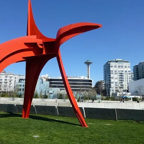The iconic red 'Eagle' sculpture by Alexander Calder in Seattle’s Olympic Sculpture Park, set against a clear blue sky. The Space Needle and modern apartment buildings are visible in the background, with green grass and a pe