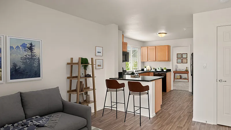 Bright, open-concept apartment featuring a cozy living room and kitchen with warm wood cabinetry. A gray sofa sits beneath framed landscape artwork, next to a wooden ladder shelf with plants and decor. The kitchen includes a breakfast bar w