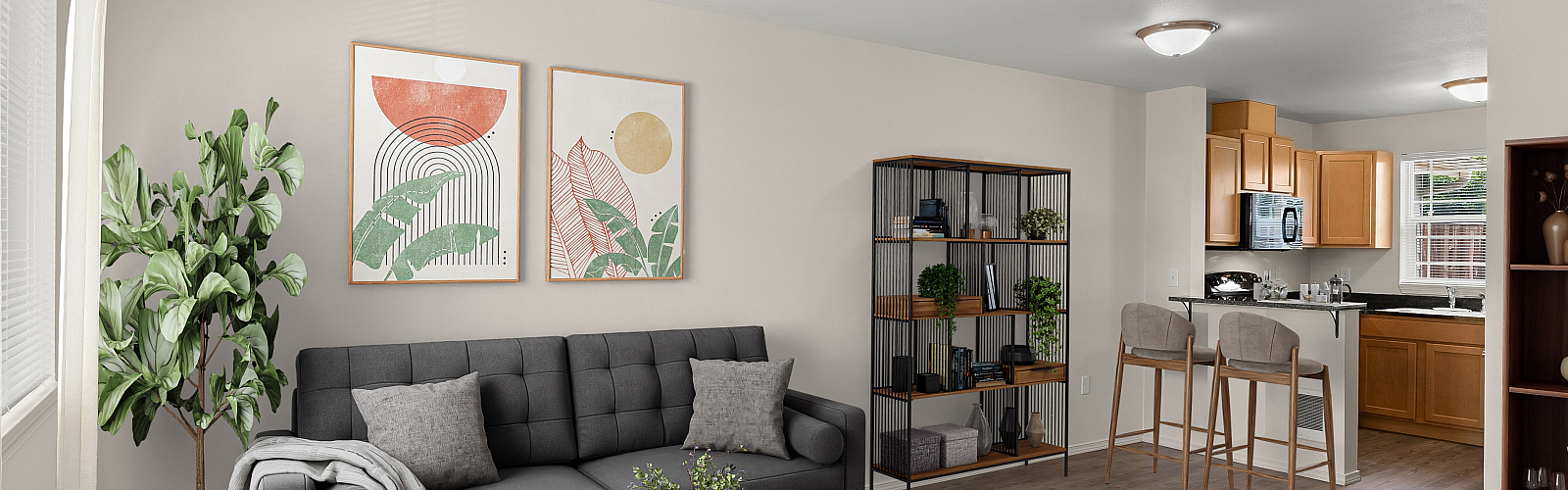 Open-concept apartment interior featuring a modern living room and kitchen. The living space includes a dark gray tufted sofa with accent pillows, a wooden coffee table, and a tall green plant. Abstract art prints hang above the couch. A ba