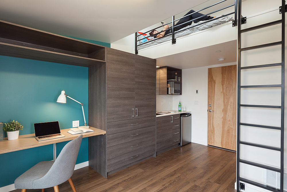Modern student apartment featuring a lofted bed with metal ladder access, built-in desk and shelving, compact kitchen with stainless steel appliances, and wood-style flooring.
