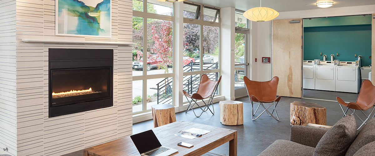 Modern apartment lounge with a cozy gas fireplace, large sectional sofa, wood coffee table, and butterfly chairs. A laundry room is visible in the background through open sliding doors, and large windows bring in natural light from outside.