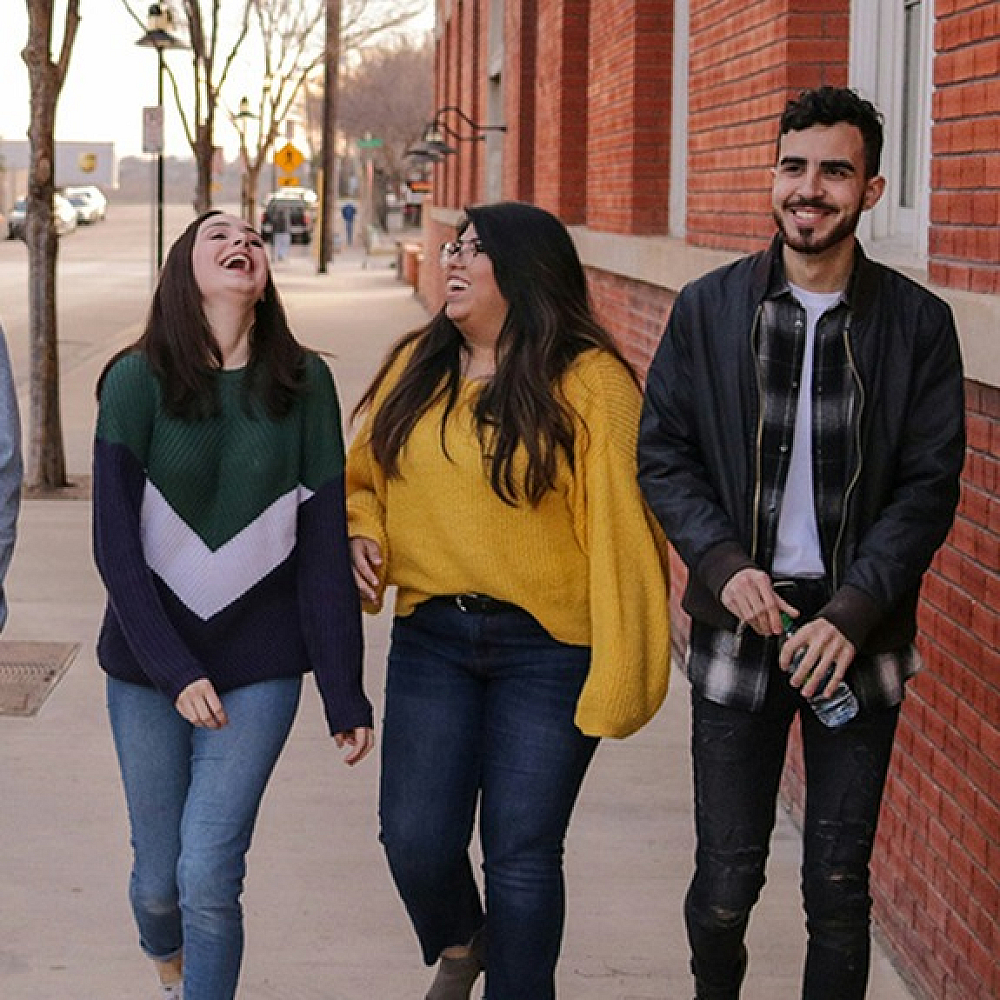 Group of five young adults walking and laughing together on a city sidewalk lined with red brick buildings, dressed casually on a sunny day.