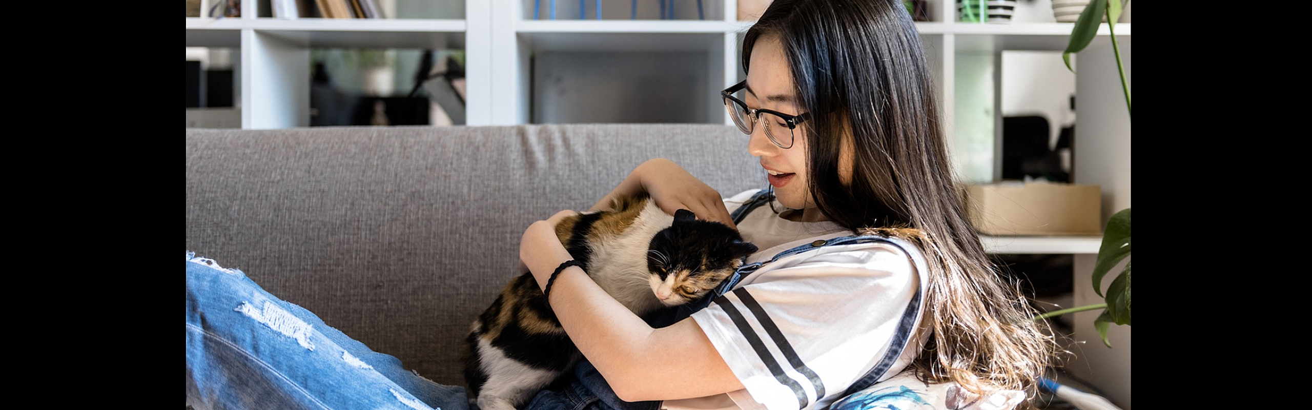 Contemporary aYoung woman lounging on a gray couch, smiling as she cuddles a calico cat in a cozy living room with books, plants, and shelves in the background.partment dining area with four black chairs around a table, hardwood floors, acc