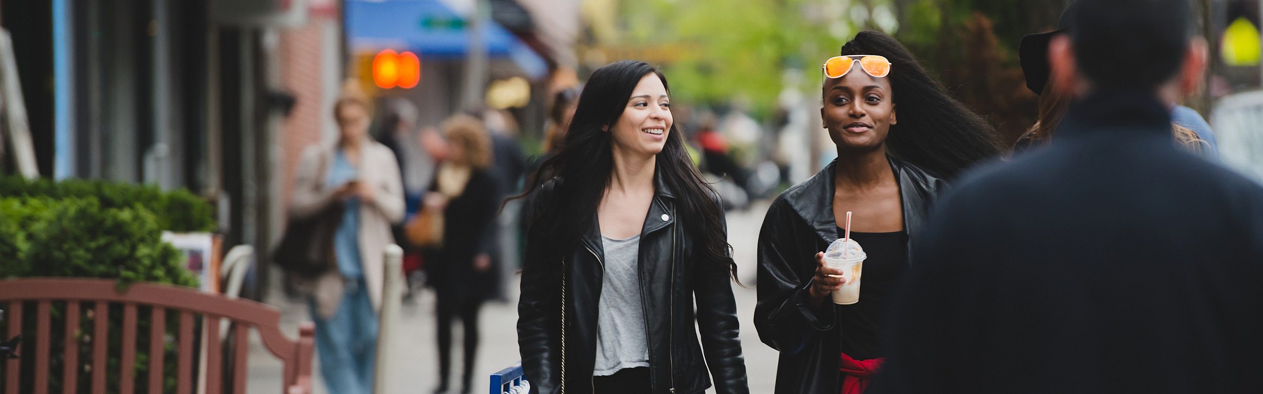 Two women walking and smiling on a busy city sidewalk, one holding a drink, surrounded by outdoor seating and pedestrians.