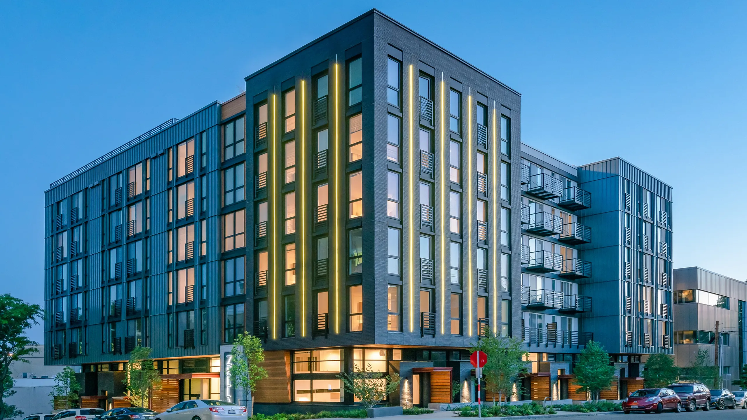 Contemporary mid-rise apartment building at dusk with glowing windows and vertical accent lighting, located on a quiet street with parked cars