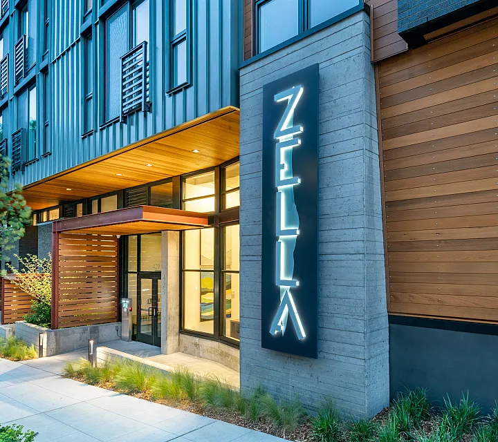 Zella apartment entrance at dusk with illuminated vertical signage, wood and metal facade, and modern landscaping