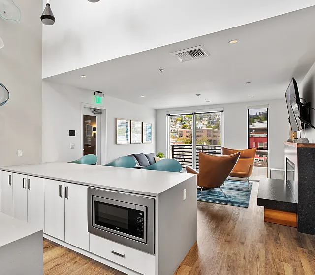 Modern apartment lounge featuring a white kitchen island with built-in microwave, stylish pendant lighting, wood-style flooring, and a cozy seating area with colorful chairs, a wall-mounted TV, and a balcony view of the city.