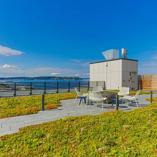 Rooftop patio with seating area and green roof garden overlooking a scenic waterfront and distant hills under a clear blue sky.