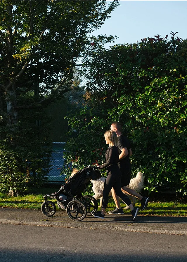 Family outdoors with a child riding a bike while parents walk with a stroller and dog on a sunny day.