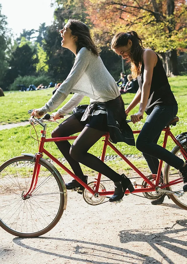 Two women riding a red tandem bicycle together through a sunny park with green grass and trees in the background.