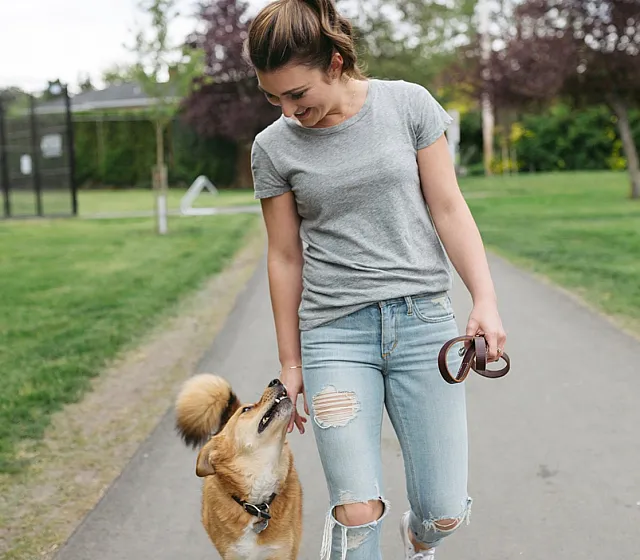 Woman walking her dog on a paved path in a park, smiling as the dog looks up at her.