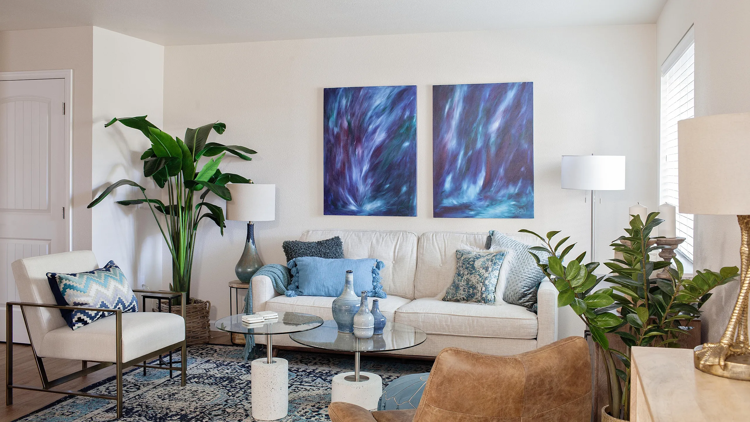 Bright living room with a white sofa, blue and patterned throw pillows, glass coffee table, modern chairs, tall green plants, and two abstract blue paintings on the wall.