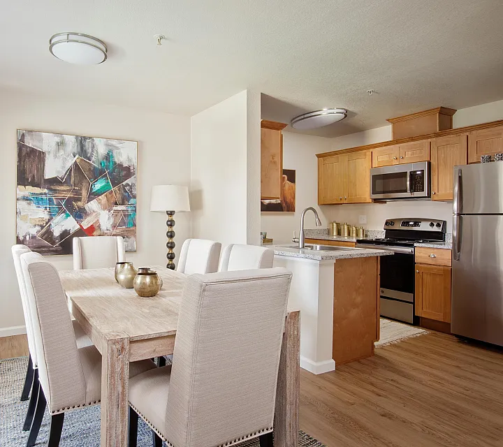 Modern apartment kitchen and dining area with wood cabinetry, stainless steel appliances, a rustic dining table, and upholstered chairs.