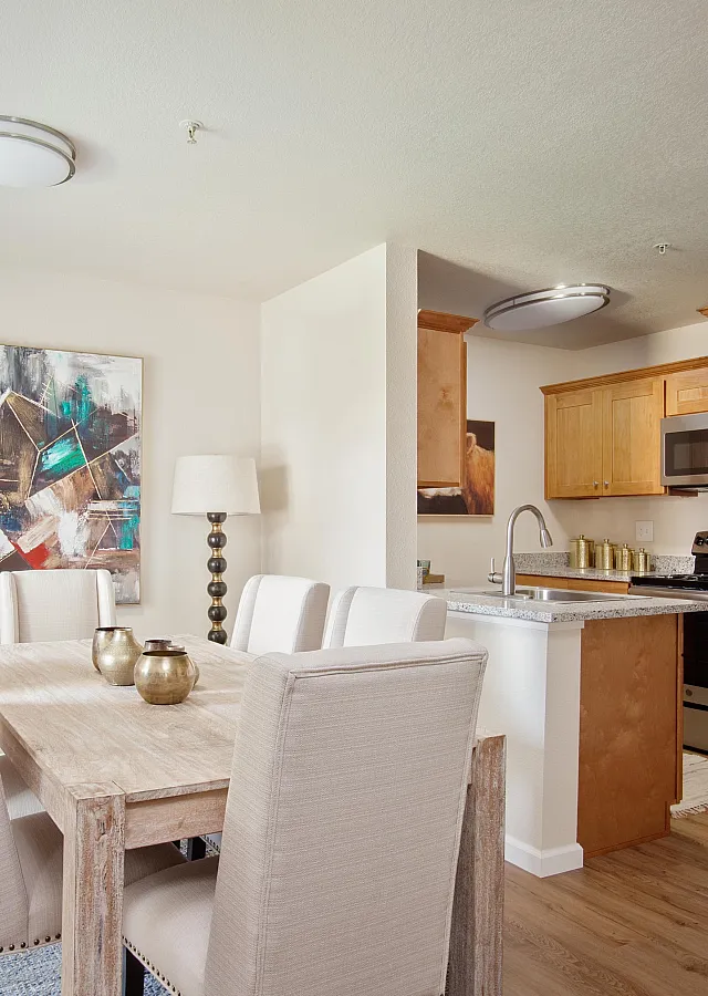 Dining area with a rustic wooden table, upholstered chairs, modern artwork, and an open kitchen with wood cabinetry and stainless steel appliances.