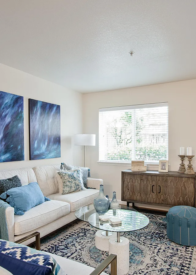 Bright living room with a cream sofa, blue accent pillows, glass coffee table, abstract wall art, and a patterned rug.