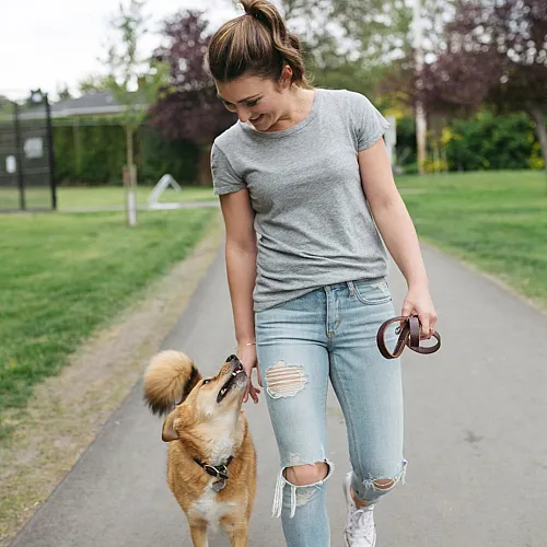 Young woman walking her dog along a paved path in a green park, smiling as the dog looks up at her.