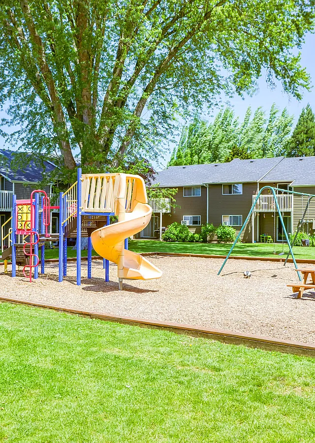 Outdoor playground with slide, swings, and climbing structures surrounded by green grass and apartment buildings.