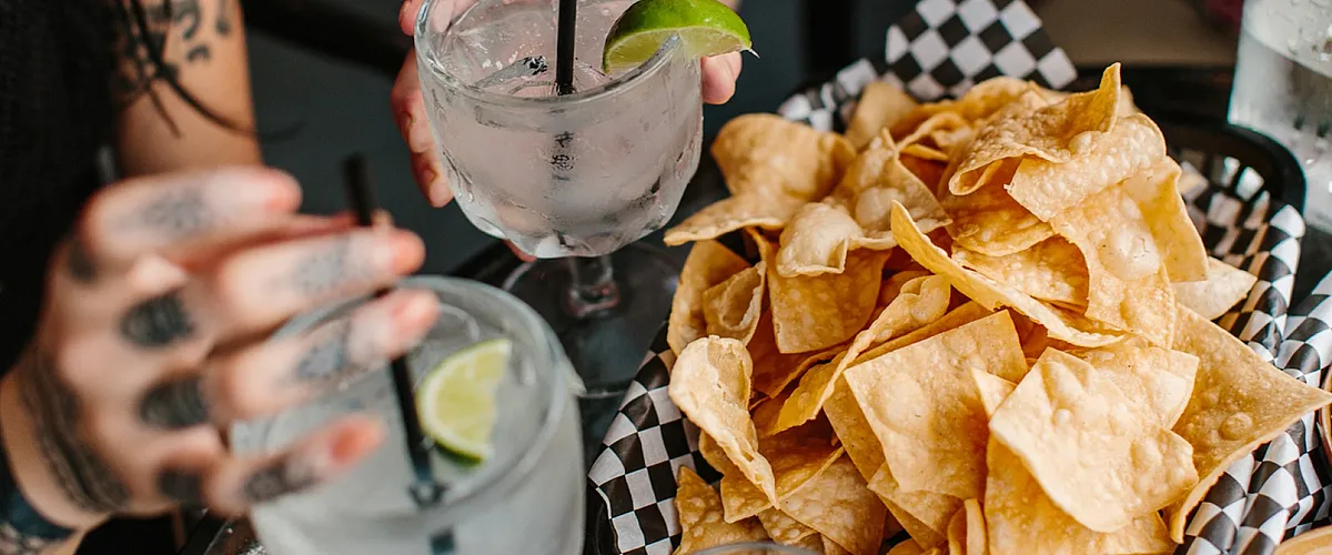 Close-up of a basket of tortilla chips next to two drinks with lime wedges.