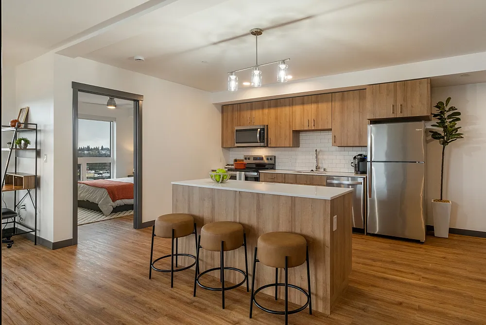 Modern apartment kitchen with wood cabinets, stainless-steel appliances, and an island with three stools, with a bedroom visible through an open doorway.