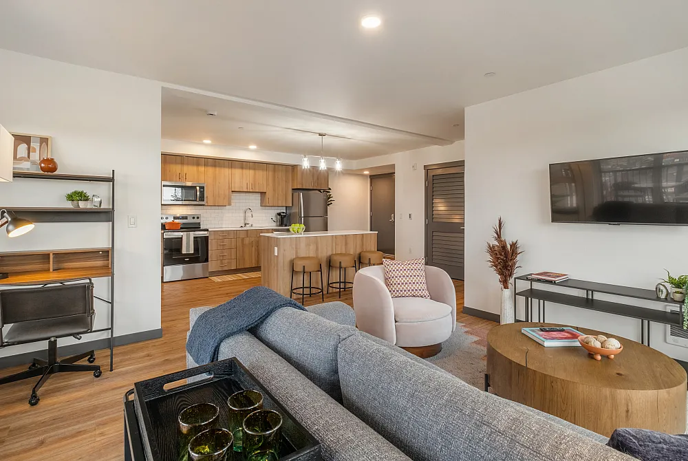Modern open-concept living room and kitchen with wood flooring, a gray sofa, mounted TV, and a kitchen island with stools.