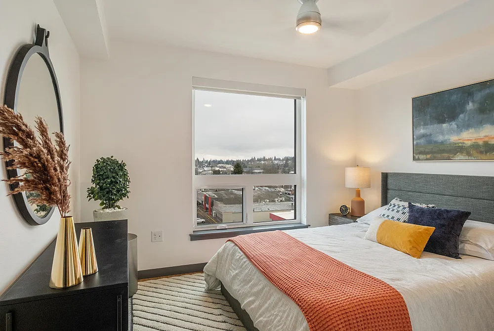 Bright bedroom with a large window, modern decor, and a bed with colorful pillows and an orange throw.