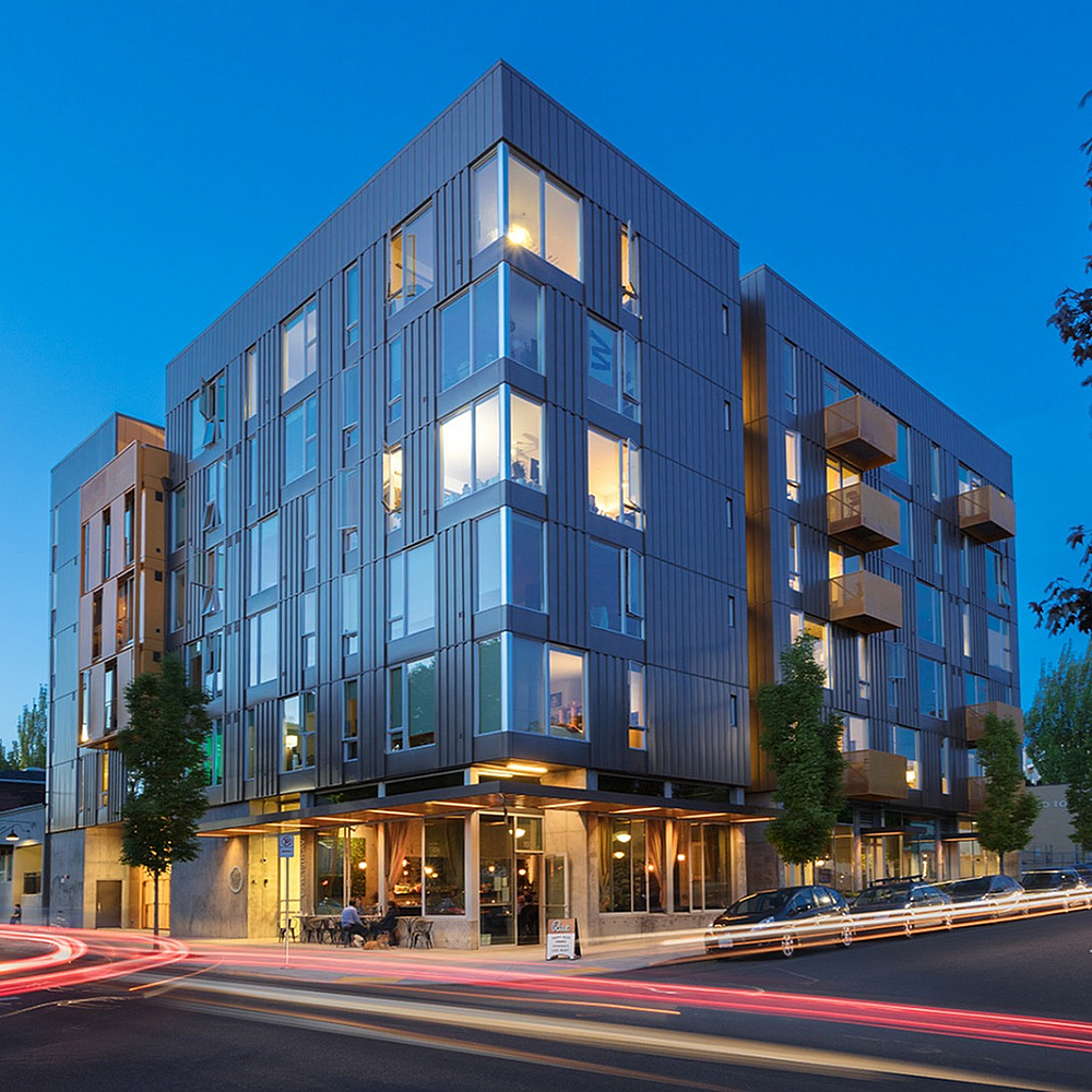 Modern mid-rise apartment building photographed at dusk with illuminated windows, street-level retail, and light trails from passing cars.
