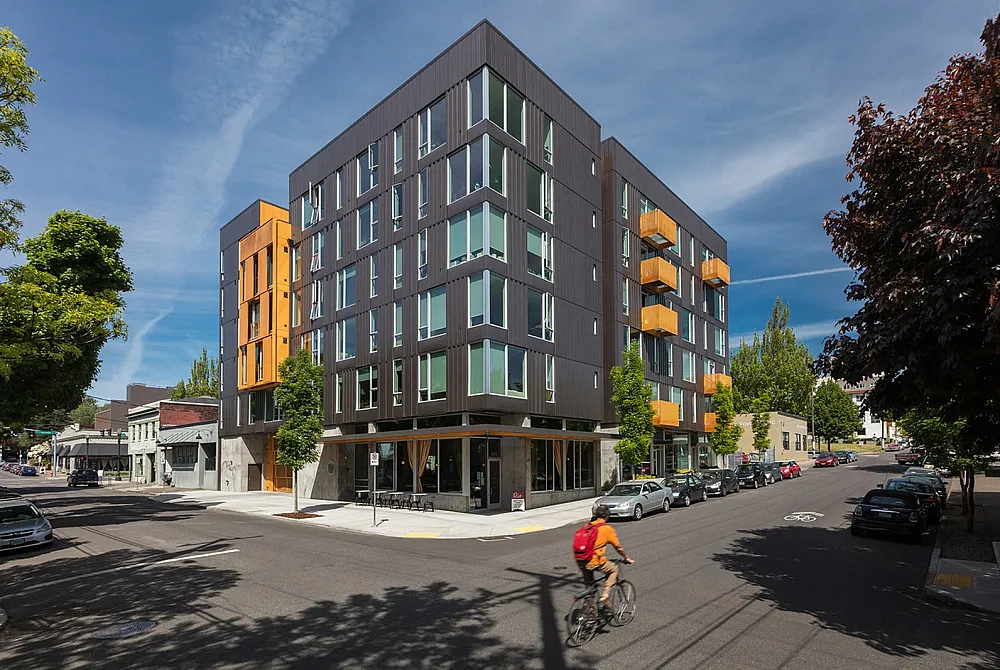 Street-level view of a modern apartment building with dark siding, bright orange accents, large windows, and a cyclist riding past on a sunny day.