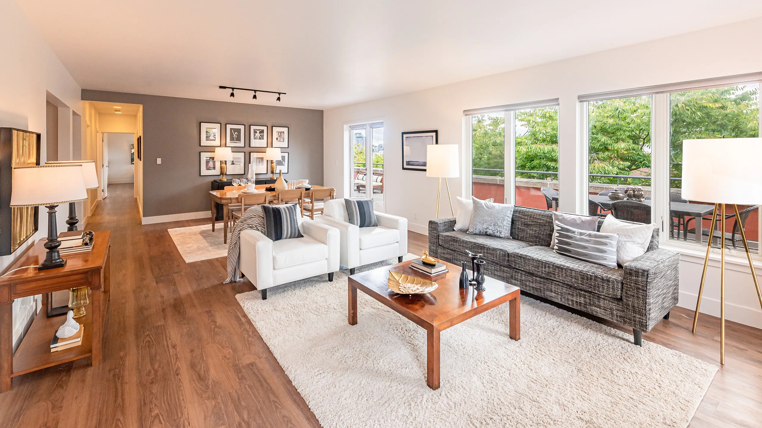 A spacious, modern living room with large windows showing greenery. Features include a beige rug, a dark gray sofa, two white armchairs, and a wooden coffee table. A dining area with a table and framed wall art is in the background, creatin