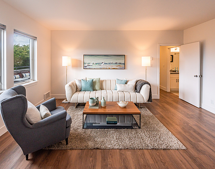 A cozy living room with a white sofa, teal and beige cushions, two lamps, and abstract wall art. A gray armchair and wooden table create a warm, inviting atmosphere.