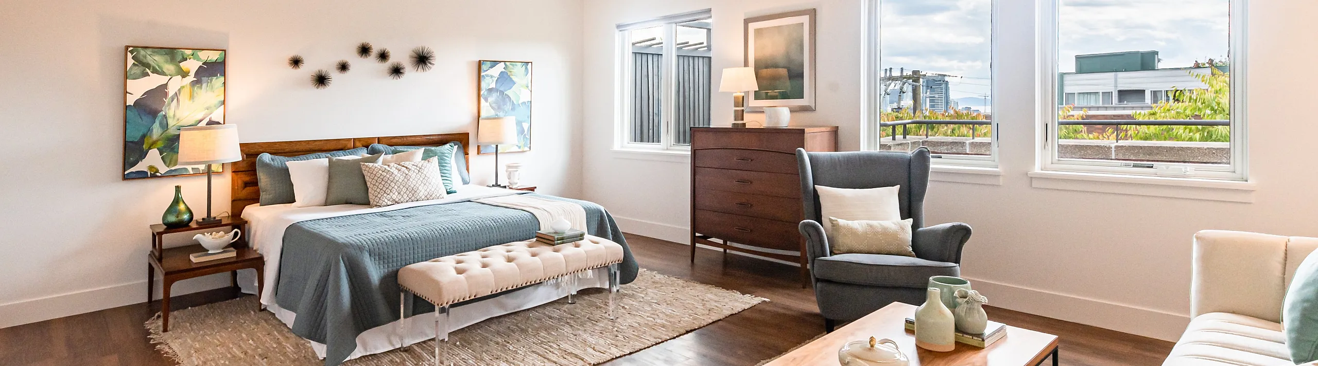 Bright bedroom with large windows, a cozy bed with blue and white linens, a gray armchair, wooden furniture, and a peaceful, inviting atmosphere.