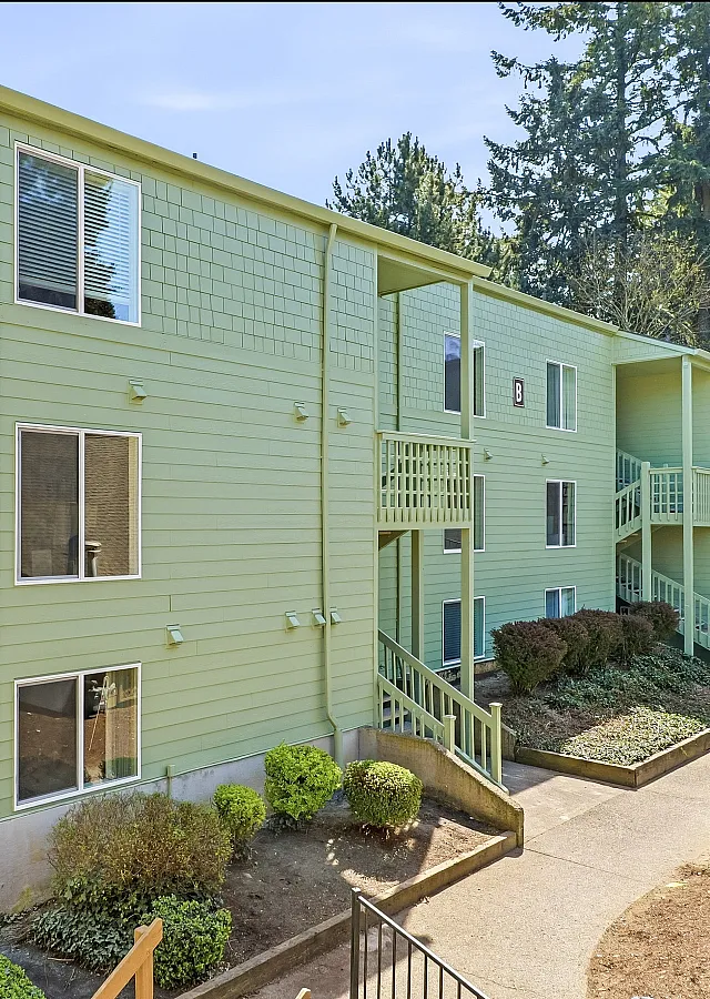 Exterior of The Finch apartments in Vancouver, WA, with tan siding, covered parking, and tall evergreen trees in the background.