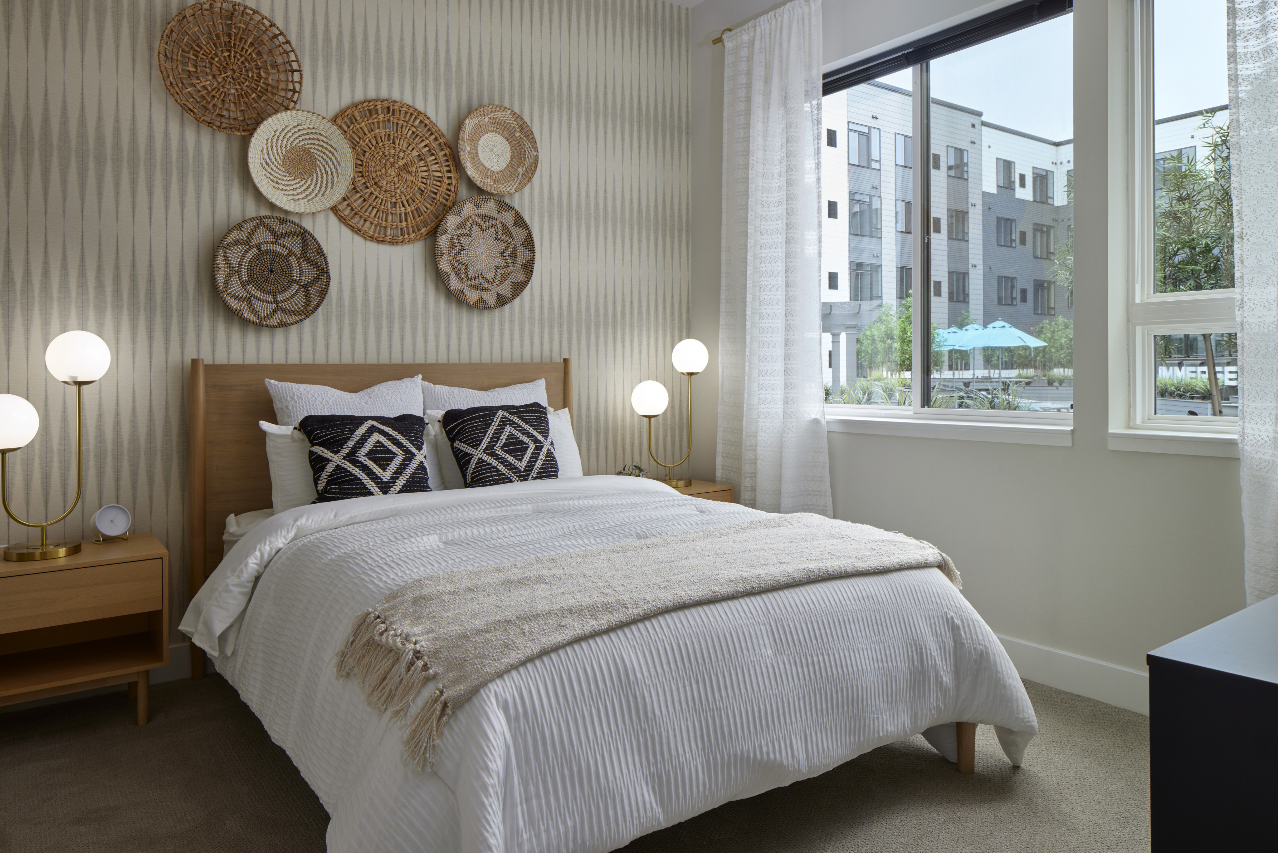 Cozy bedroom with a wooden bed, white bedding, woven wall decor, and a large window overlooking a pool.