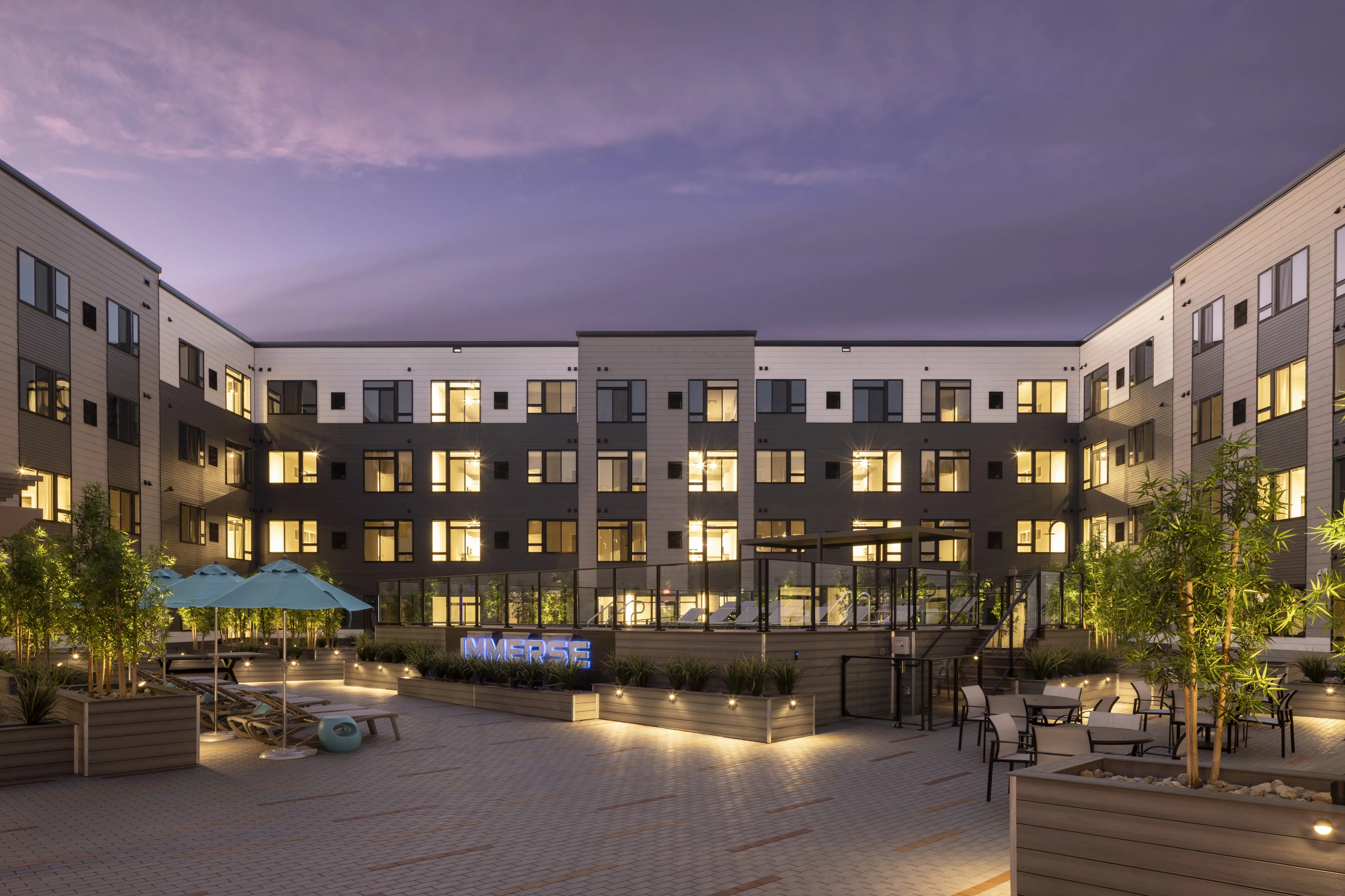 Modern apartment courtyard at Luxor Montgomeryville with lounge chairs, umbrellas, greenery, and a pool, lit under a twilight sky.