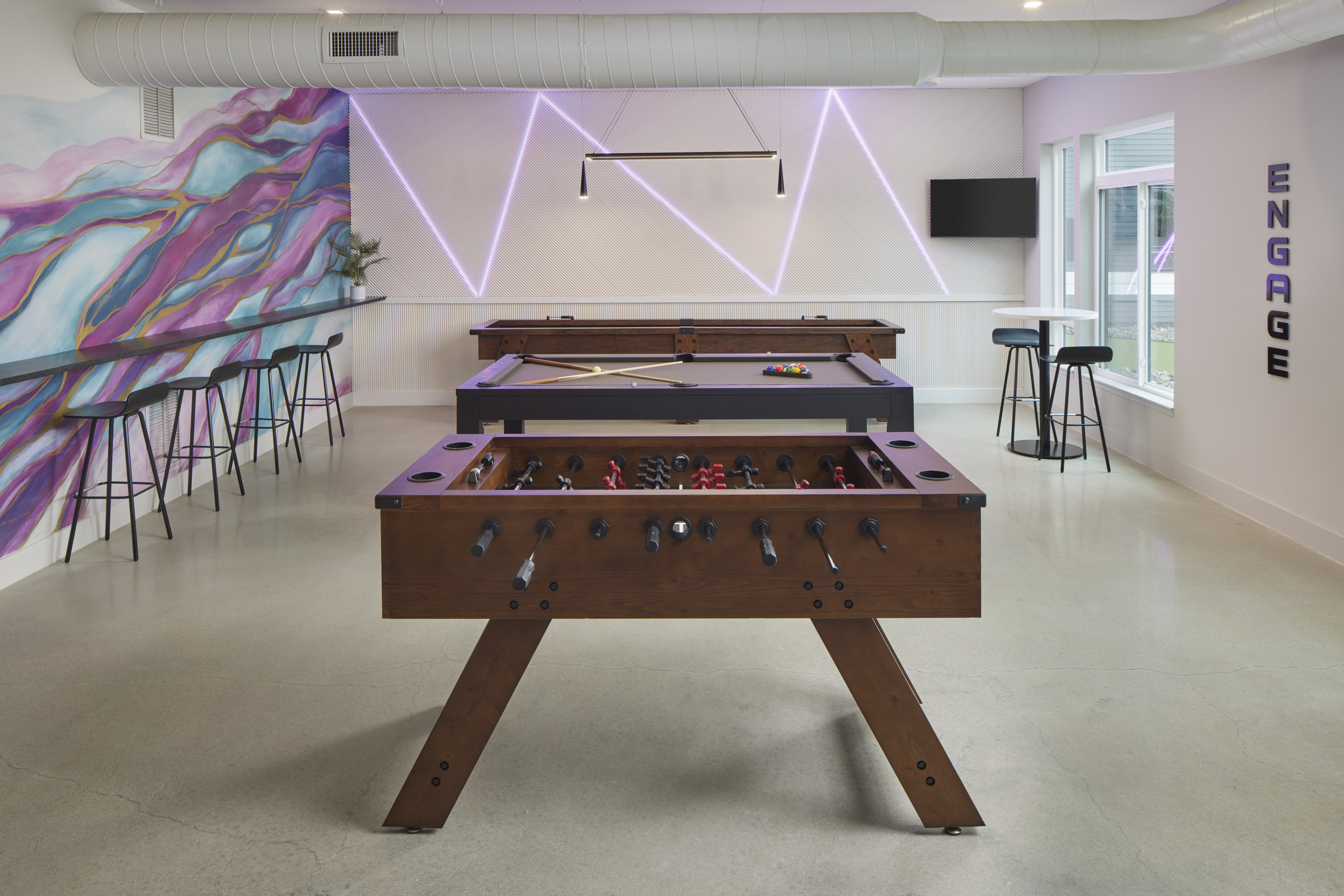 Modern community game room with foosball and pool tables, bar seating, neon wall lights, and a colorful mural.