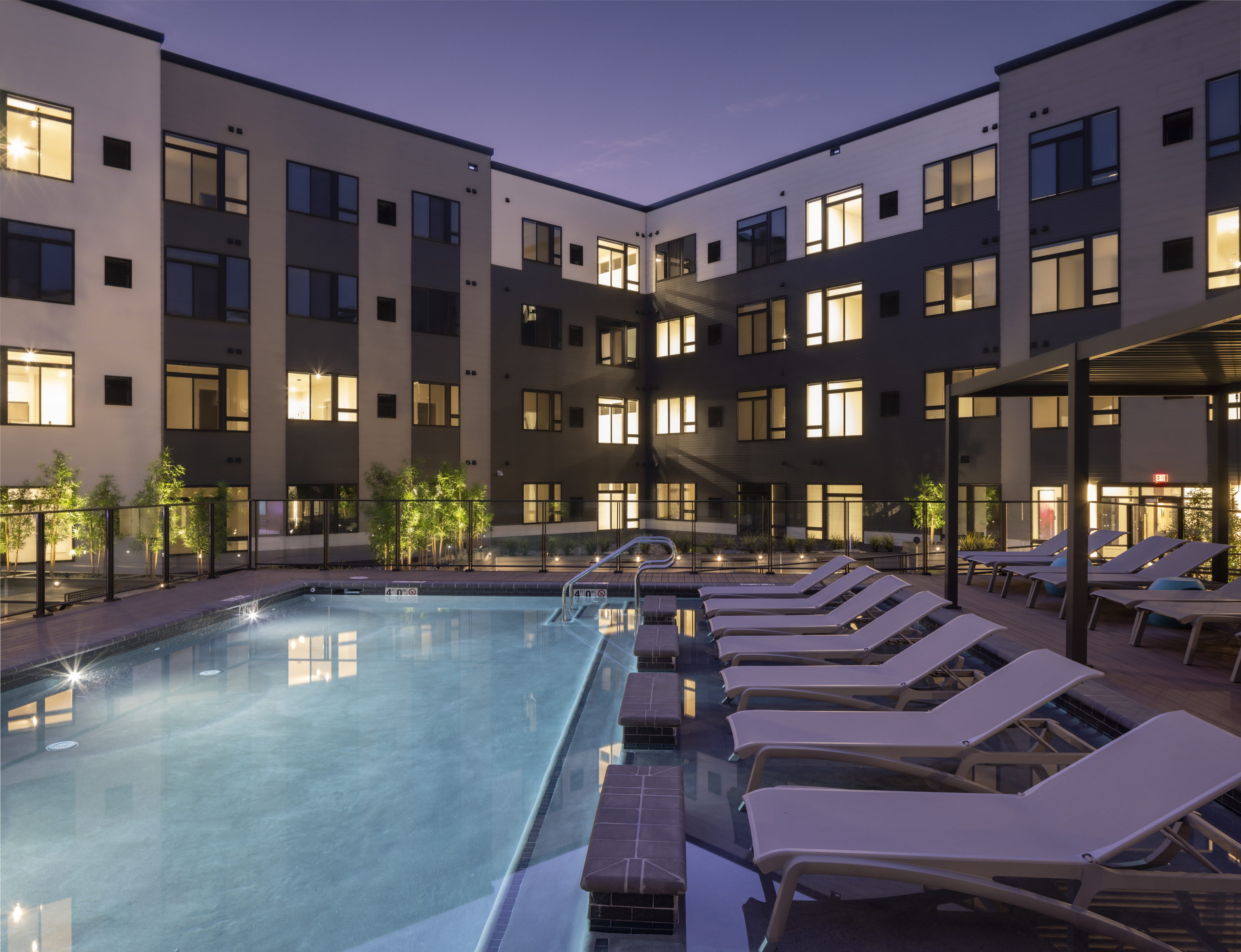 Luxurious pool area at night with lounge chairs, warm apartment lighting, and modern glass fencing.