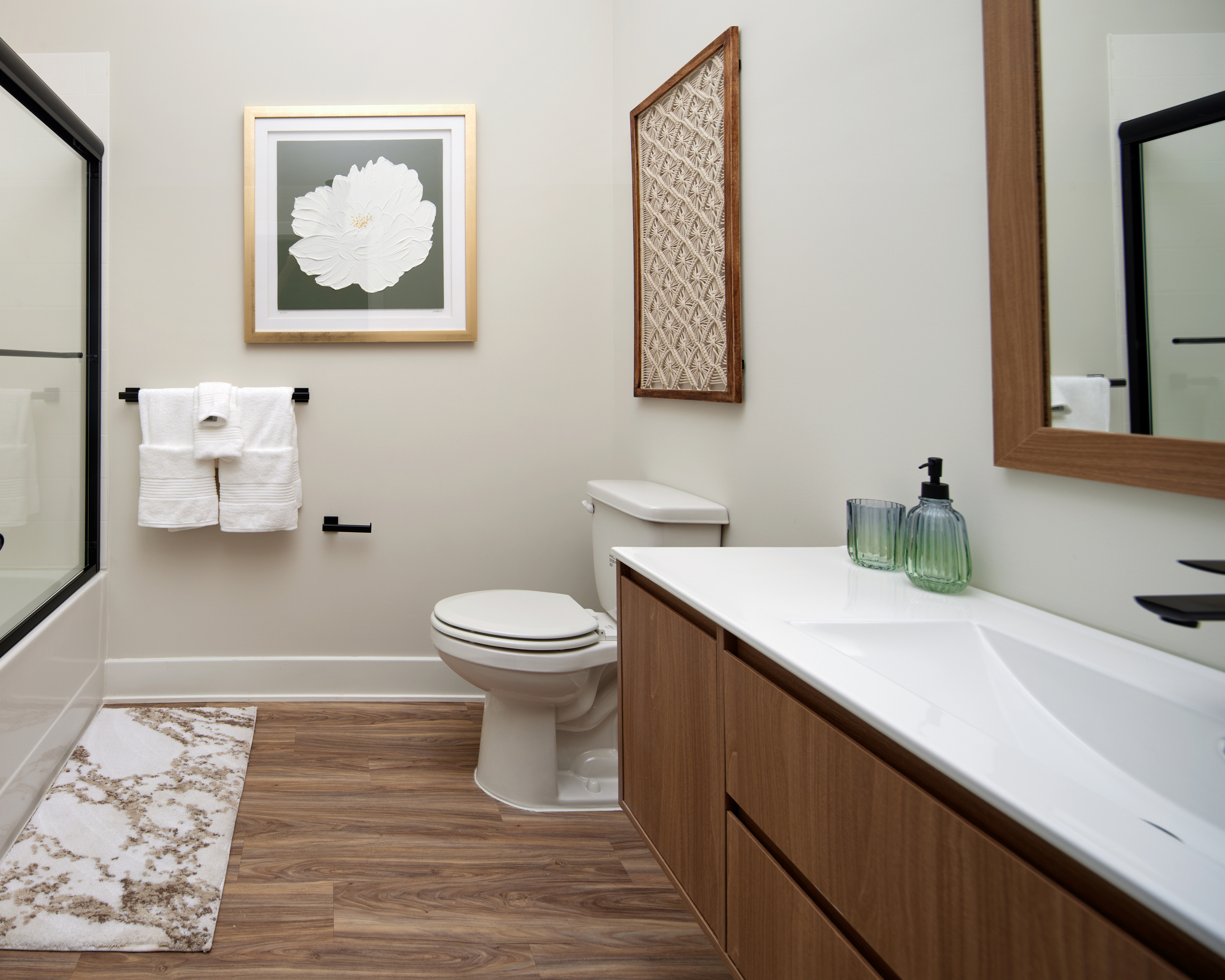 Modern apartment bathroom with wood accents, white countertop, glass shower, and decorative wall art.