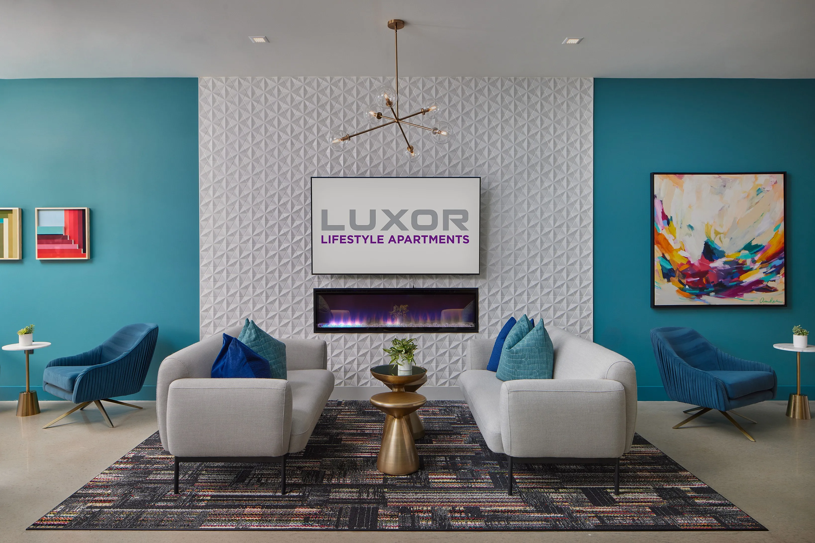 Luxor Lifestyle Apartments lounge area showcasing modern furniture, vibrant artwork, and a sleek wall fireplace in a stylish teal and white setting.